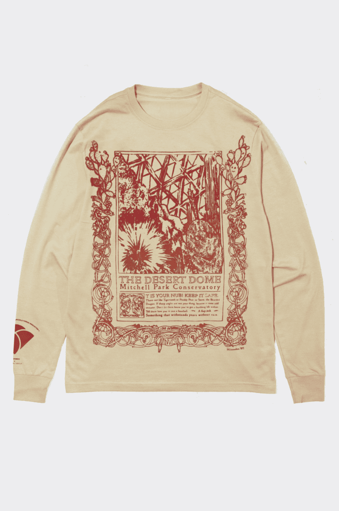 A tan long-sleeve shirt with a vintage-style, art nouveau inspired illustration and text in red and orange. The design features various cacti, including one with a smiley face, a geodesic pattern in the background, and ornate borders, with the words "The Tropical Dome" and "Milwaukee Mitchell Park Conservatory". Below is a poem from Anja Notanja Sieger, reading: "It is your nub! Keep it safe. Thorn out like Tigertooth or Prickly Pear, or Steve, the Bearded Dragon. If sharp angles are not your thing, become a stone cold imitator. Don't let them know you've got a burbling life within. Tell them how you're just a baseball. A flapjack. Something that withstands years without rain."