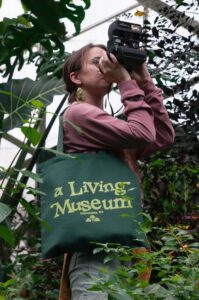 A person holding a camera is taking a photo in a greenhouse. They are carrying a green tote bag with "a Living Museum" written on it. The background is filled with various plants.