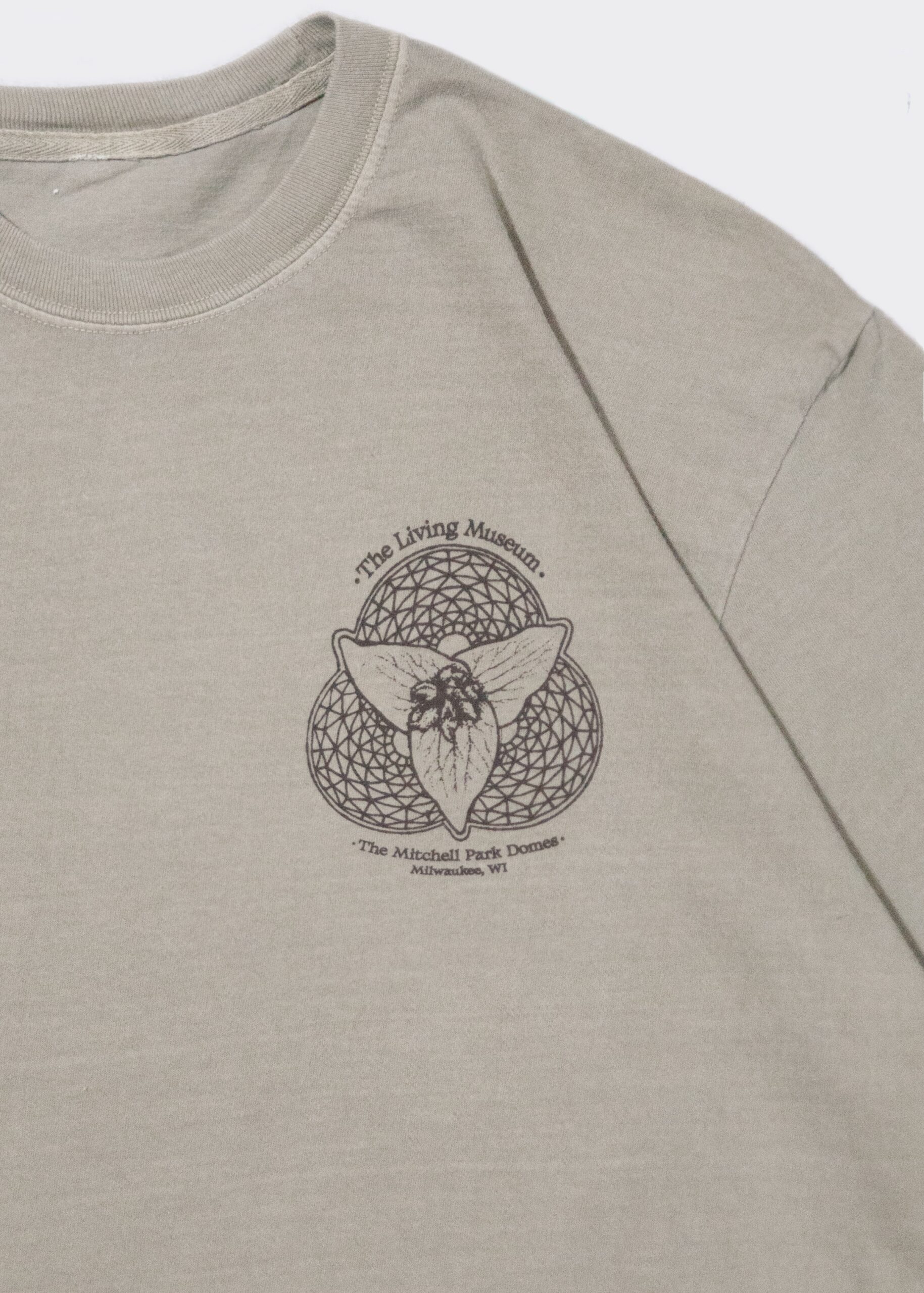 A sage green T-shirt features a printed design with the text "The Living Museum" above an illustration of an insect combined with flowers. Below the design, it reads "The Mitchell Park Domes, Milwaukee, WI." The illustration is enclosed within a detailed geometric pattern.