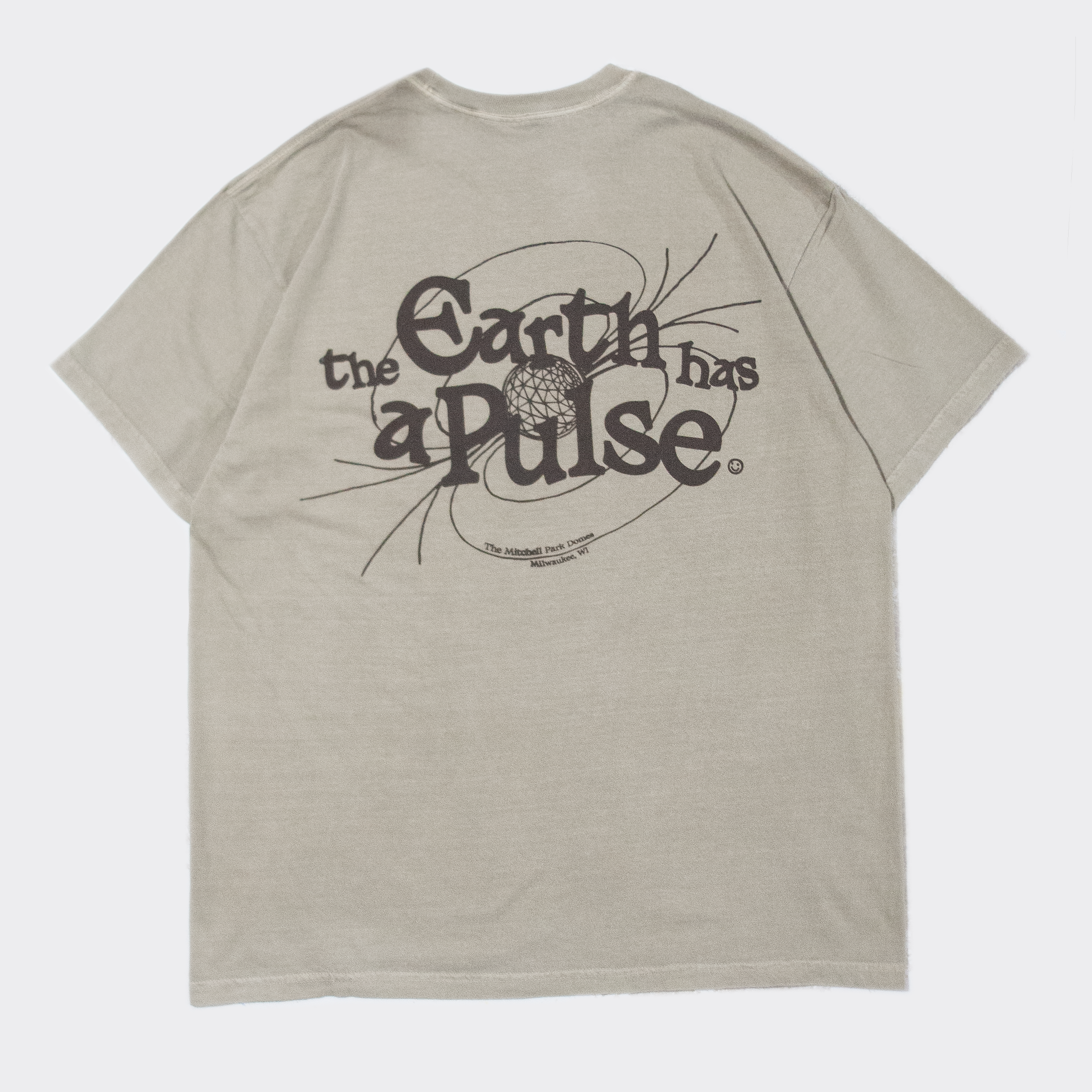 A sage-green t-shirt with brown text and design on the back. The text reads, "the Earth has a pulse" with a surrounding illustration resembling an orbit and a globe at the center. Additional smaller text below the design reads "Mitchell Park Domes / Milwaukee, WI".