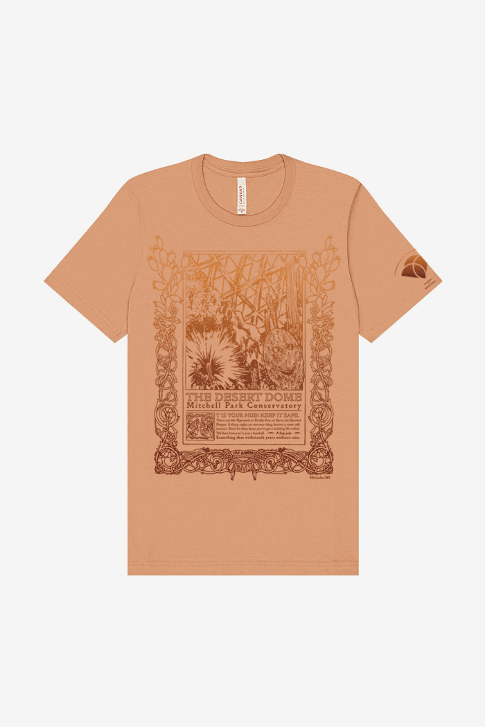 A tan short-sleeve shirt with a vintage-style, art nouveau inspired illustration and text in red and orange. The design features various cacti, including one with a smiley face, a geodesic pattern in the background, and ornate borders, with the words "The Tropical Dome" and "Milwaukee Mitchell Park Conservatory". Below is a poem from Anja Notanja Sieger, reading: "It is your nub! Keep it safe. Thorn out like Tigertooth or Prickly Pear, or Steve, the Bearded Dragon. If sharp angles are not your thing, become a stone cold imitator. Don't let them know you've got a burbling life within. Tell them how you're just a baseball. A flapjack. Something that withstands years without rain."