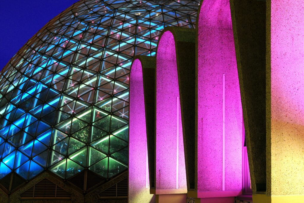 The Mitchell Park Domes and their arched entrance illuminated in blue, pink, and gold at night